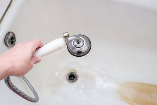 hand holding shower head with running water against rusty bathtube with limescale