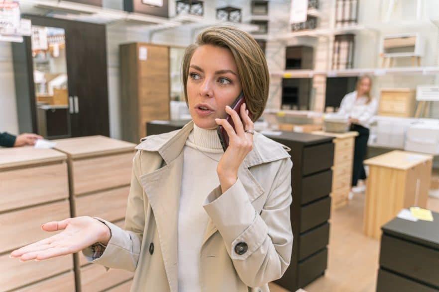 Woman in Beige Coat Talking on Phone in Furniture Shop